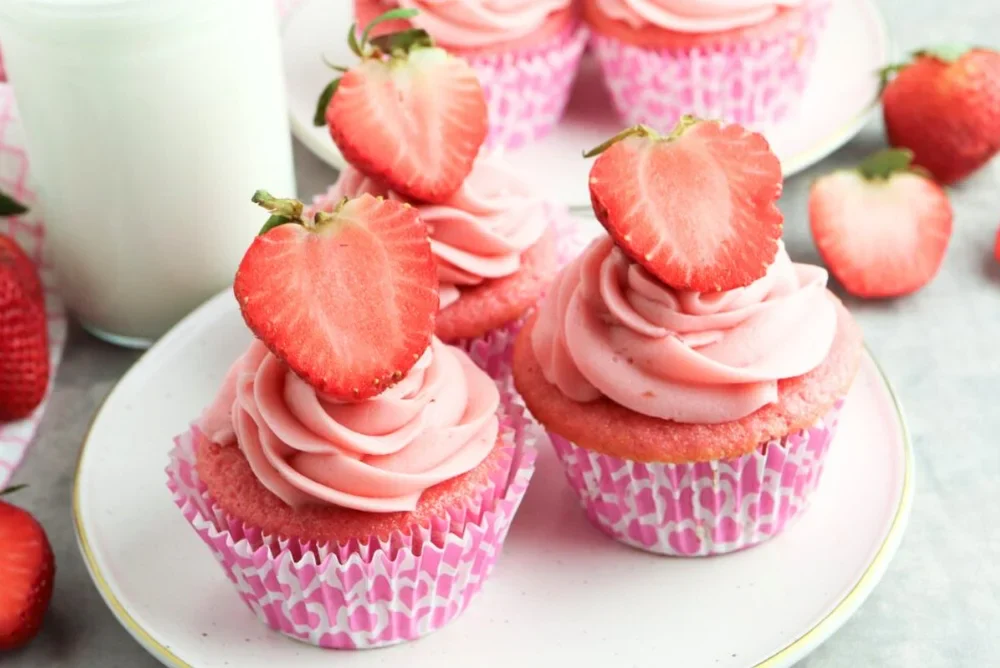 Pink frosted cupcakes topped with halved strawberries on a white plate, with whole strawberries and a glass of milk in the background.