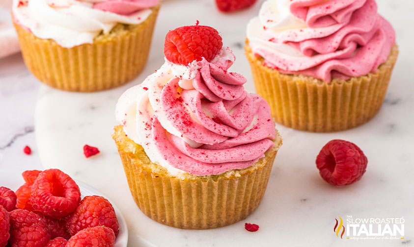 Three cupcakes with pink and white swirled frosting, each topped with a raspberry, are arranged on a marble surface next to a small pile of fresh raspberries.