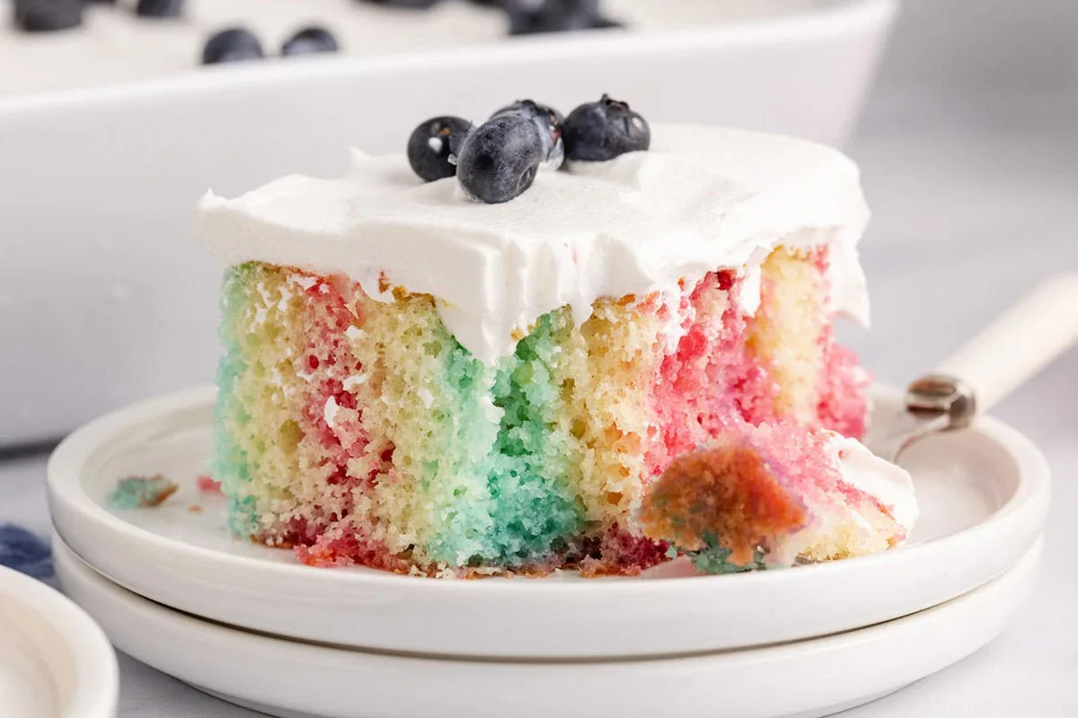 A slice of colorful poke cake with white frosting, topped with blueberries, sits on a plate with a forkful taken out.