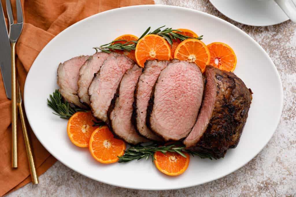 Sliced roast beef arranged on a white platter, garnished with fresh rosemary sprigs and sliced oranges.