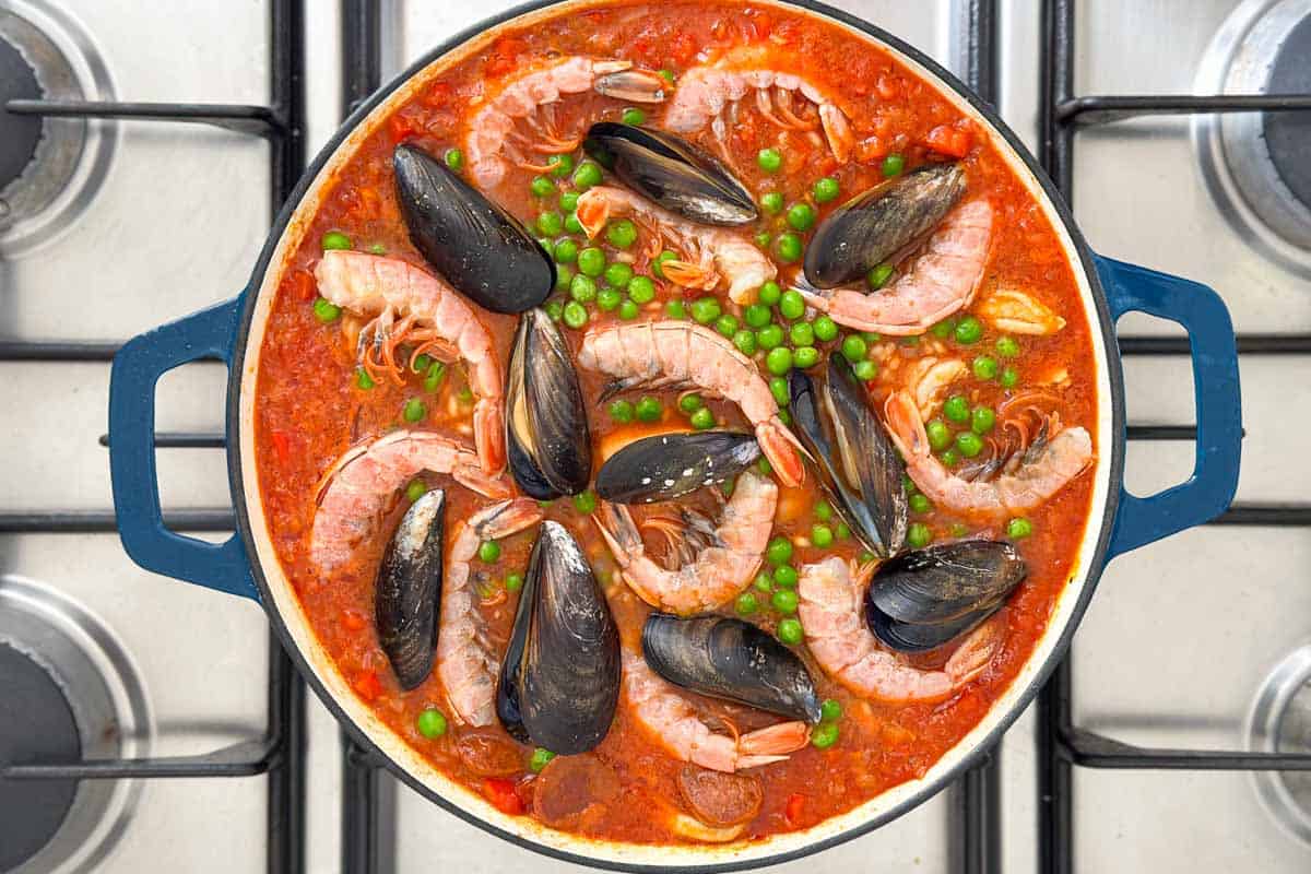 A pot of seafood stew with shrimp, mussels, green peas, and tomato sauce sits on a stovetop.