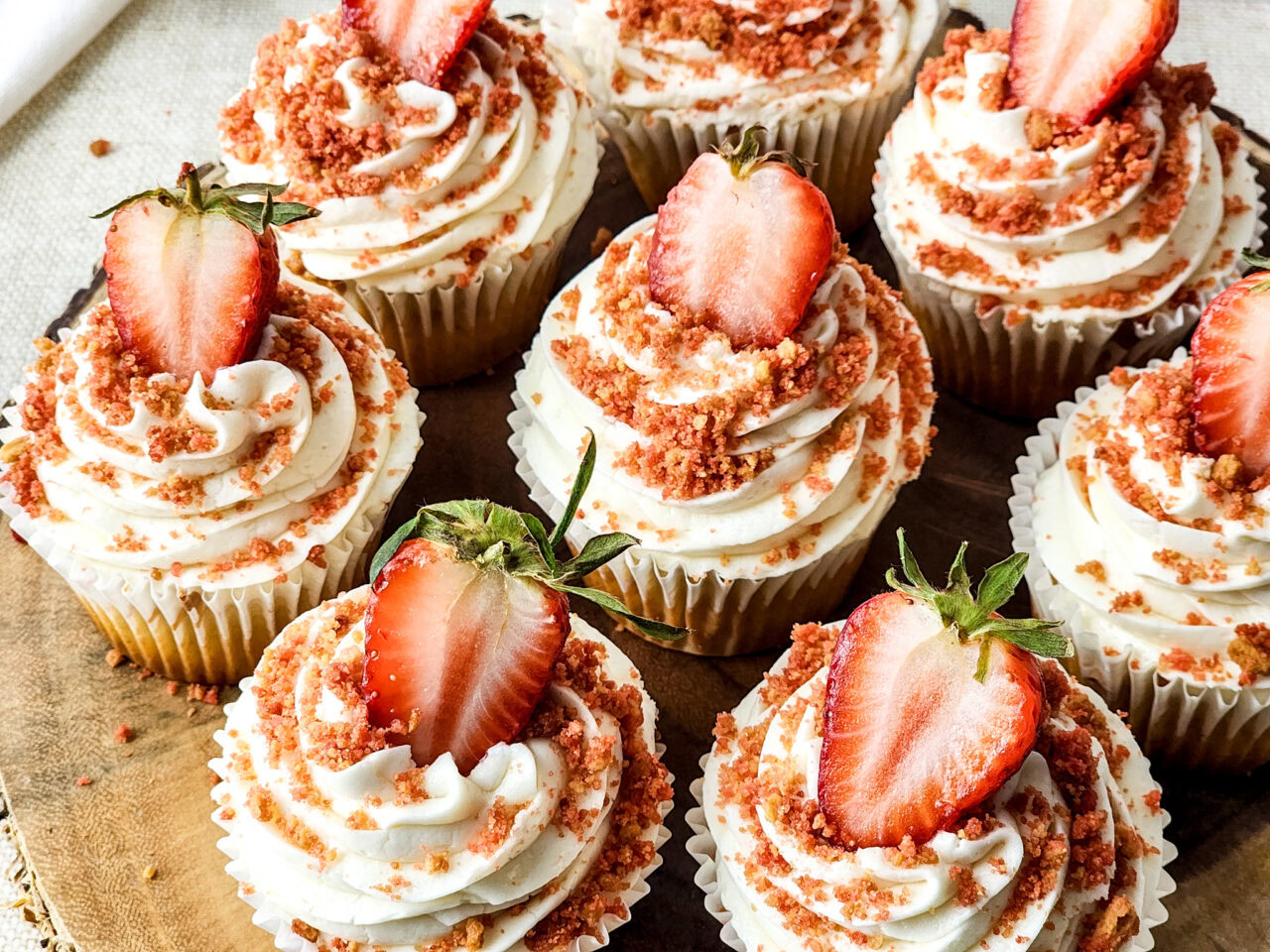 Eight cupcakes with swirled white frosting, strawberry crumbs, and half a fresh strawberry on top, arranged on a wooden surface.