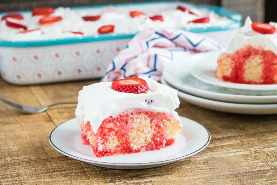 A slice of strawberry poke cake topped with whipped cream and a strawberry slice, served on a plate with the rest of the cake in a baking dish in the background.