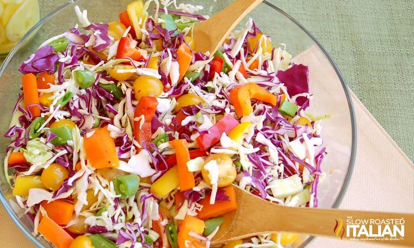 A glass bowl of colorful chopped salad with red and green cabbage, cherry tomatoes, and bell peppers, mixed with wooden salad servers.