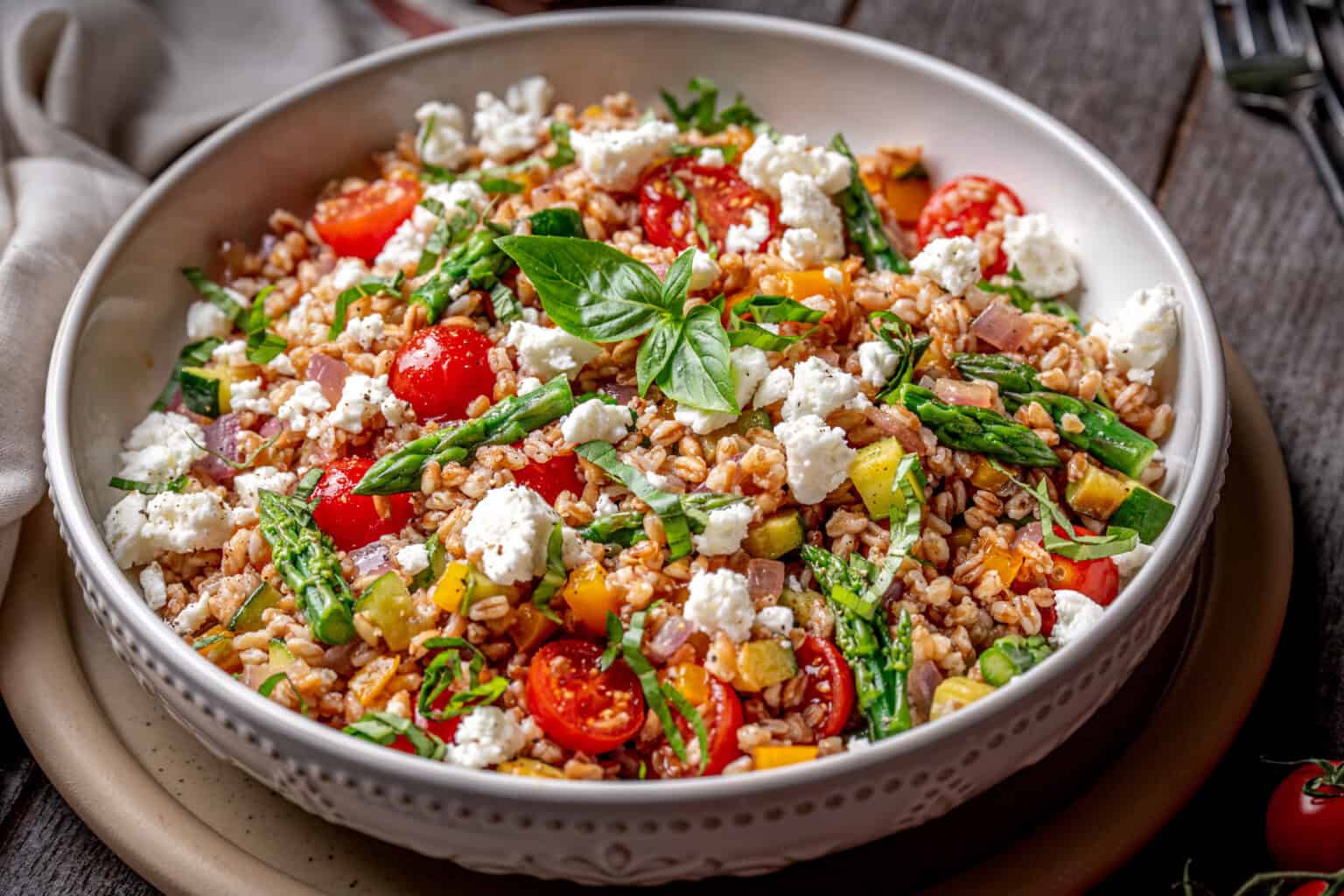 A bowl of farro salad with cherry tomatoes, asparagus, diced yellow peppers, red onions, crumbled feta cheese, and fresh basil on top.