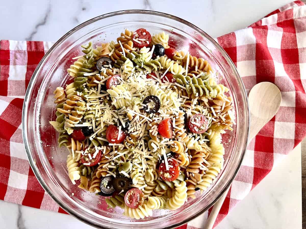 A glass bowl of tri-color rotini pasta salad with cherry tomatoes, black olives, and shredded cheese on a red and white checkered cloth.