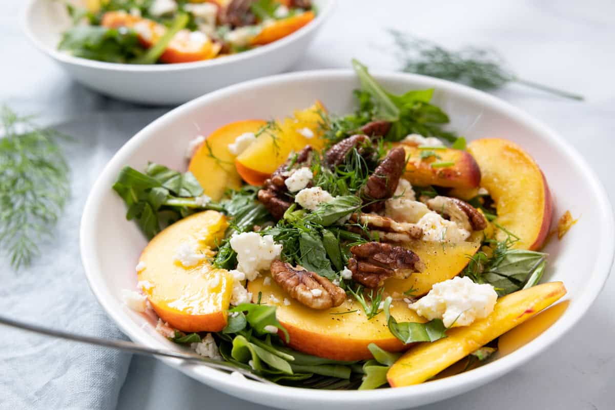 A bowl of salad with arugula, peach slices, pecans, crumbled cheese, and fresh dill on a light surface.