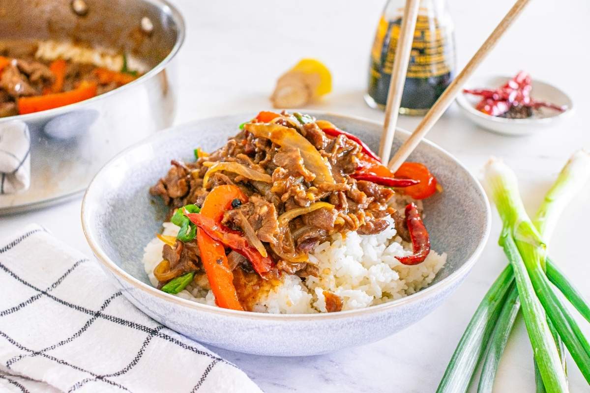 A bowl of white rice topped with stir-fried beef, red bell peppers, and onions, with chopsticks resting on the bowl. Green onions and a sauce bottle are nearby.