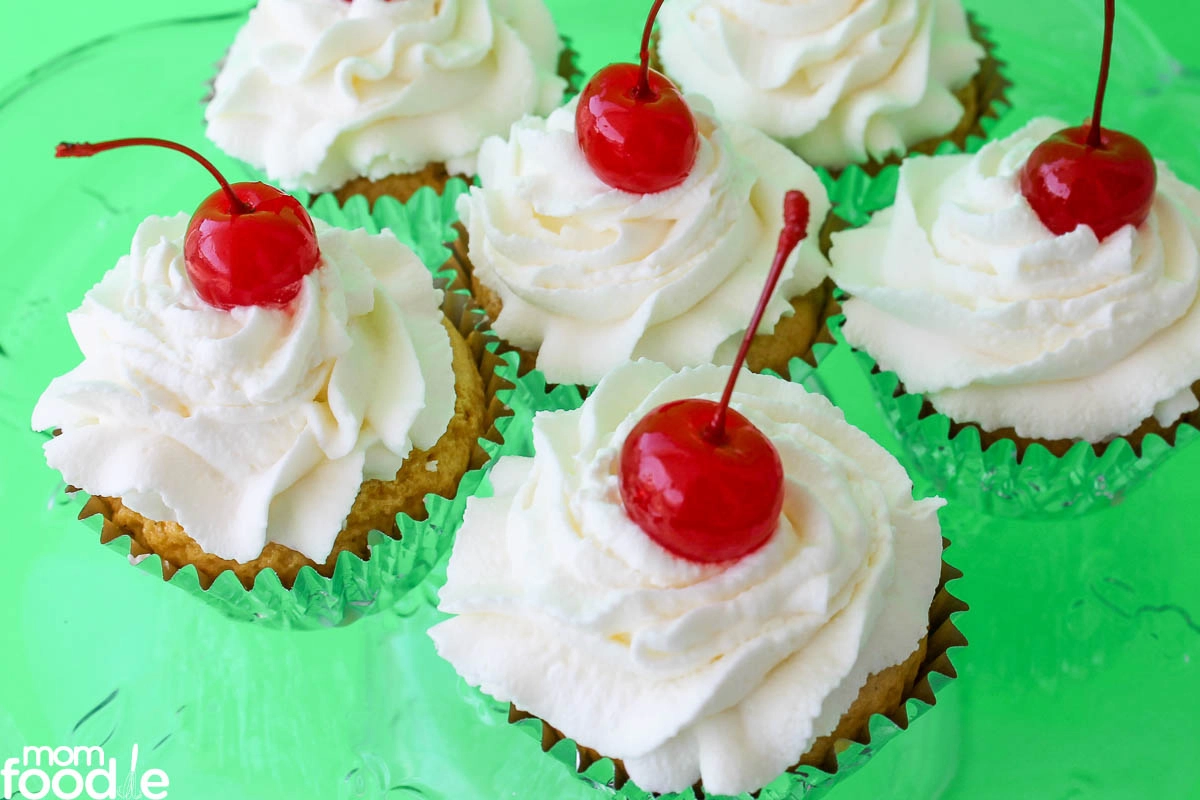 Six cupcakes with swirled white frosting and topped with maraschino cherries arranged on a green surface.