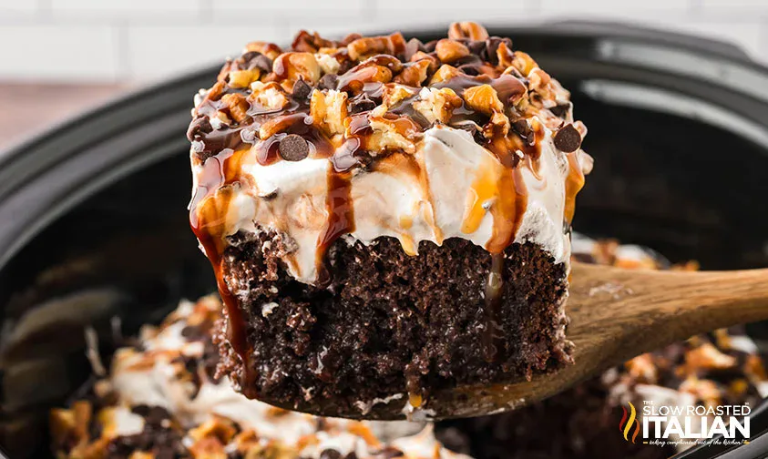 A slice of chocolate cake topped with whipped cream, chopped nuts, chocolate chips, and caramel sauce, being lifted with a wooden spatula.