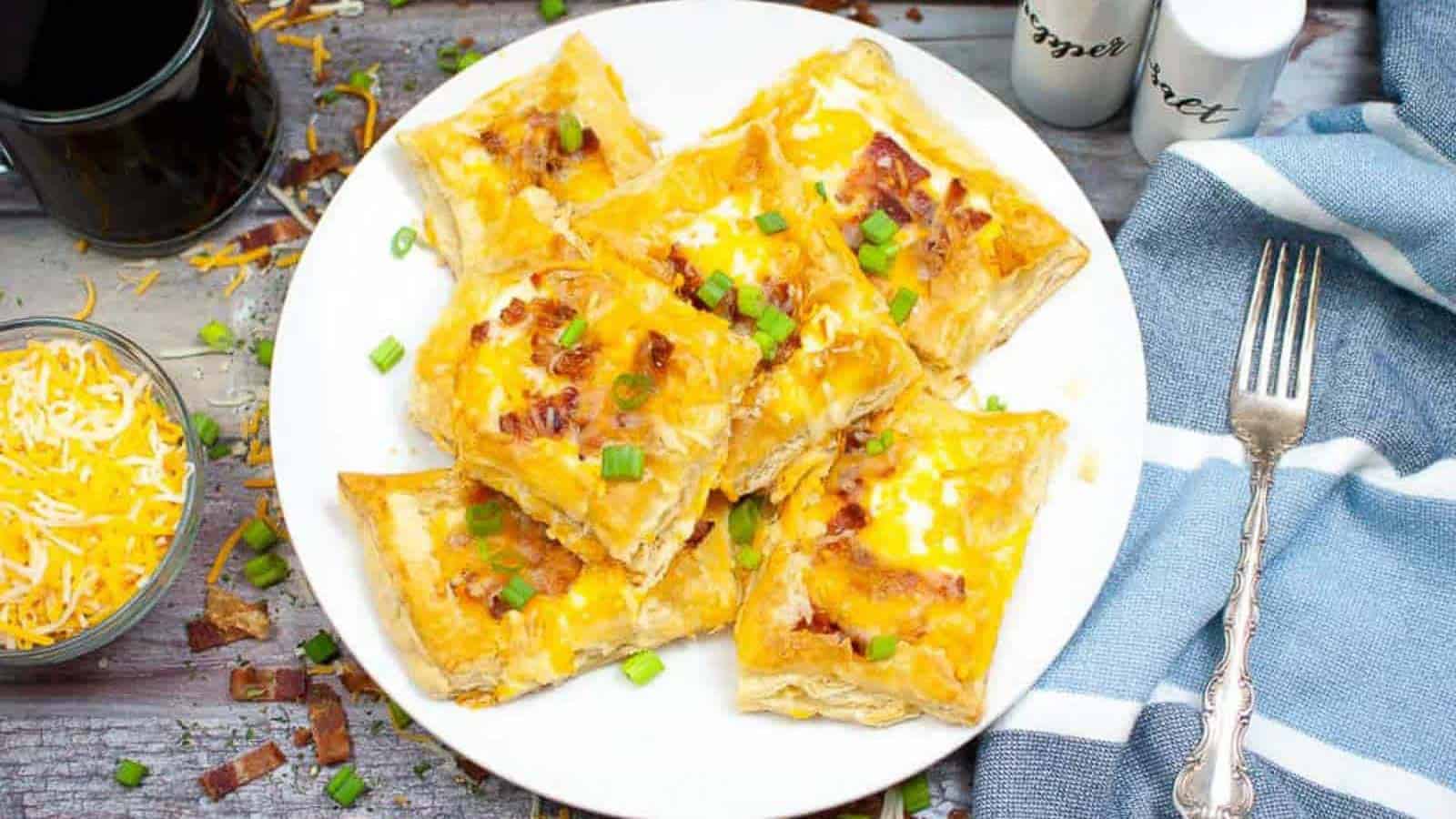 A white plate with six puff pastry squares topped with melted cheese, bacon bits, and chopped green onions, placed on a wooden table with a fork, napkin, and shredded cheese nearby.