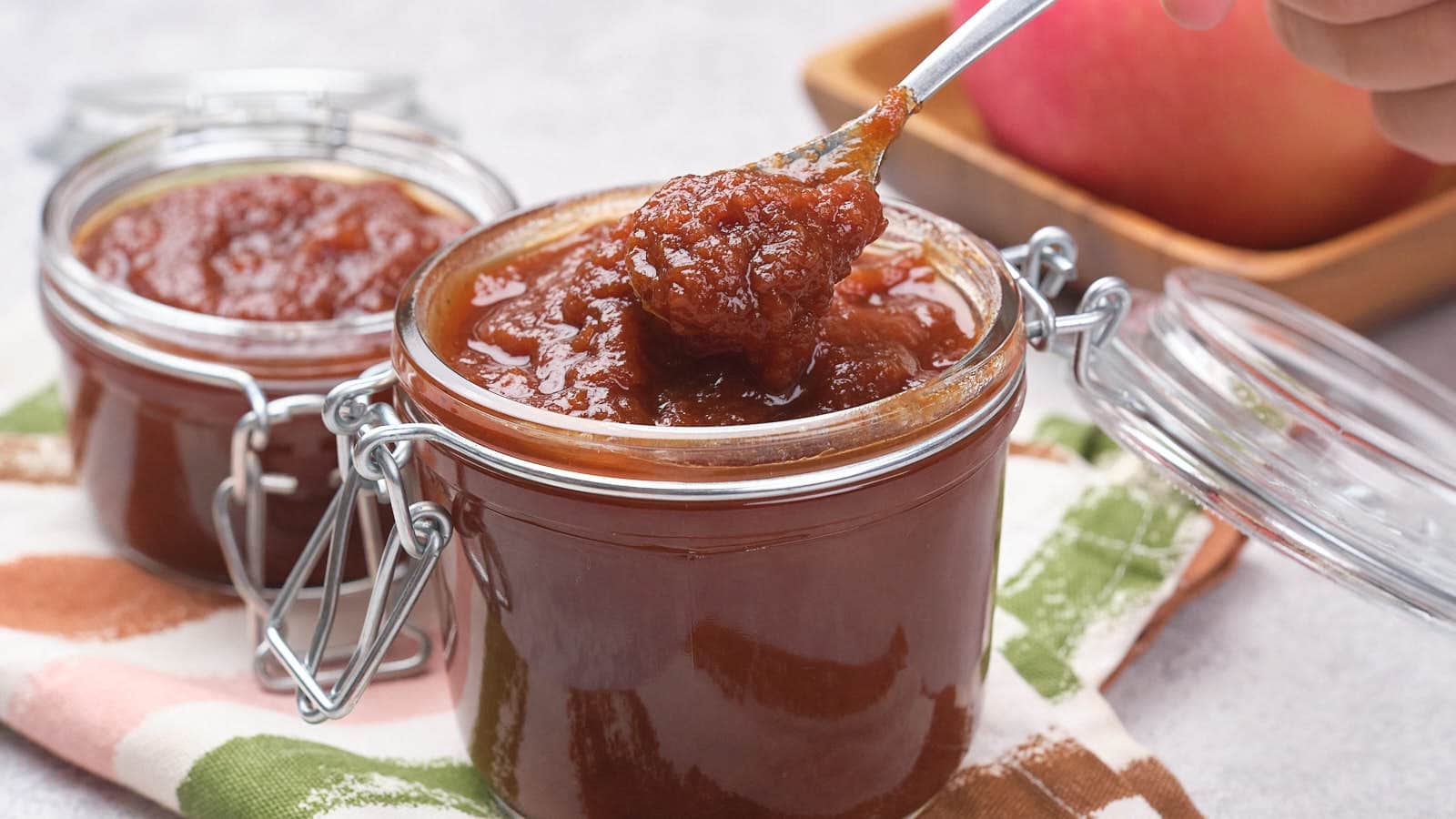 A spoon scoops thick, brown apple butter from a glass jar with a hinged lid; another jar and an apple are in the background.