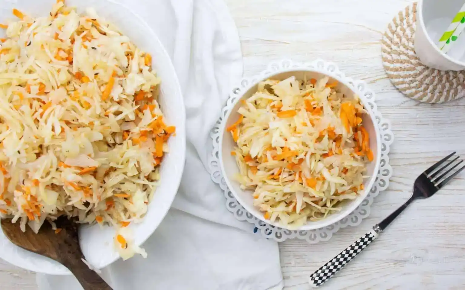 Two bowls of shredded cabbage and carrot salad are placed on a white table with a fork, napkin, and a cup.
