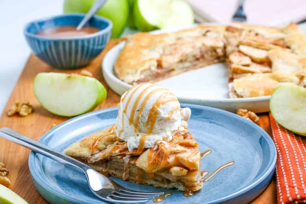 A slice of apple pie topped with vanilla ice cream and caramel sauce sits on a blue plate, with a fork beside it; a whole pie and green apple slices are in the background.