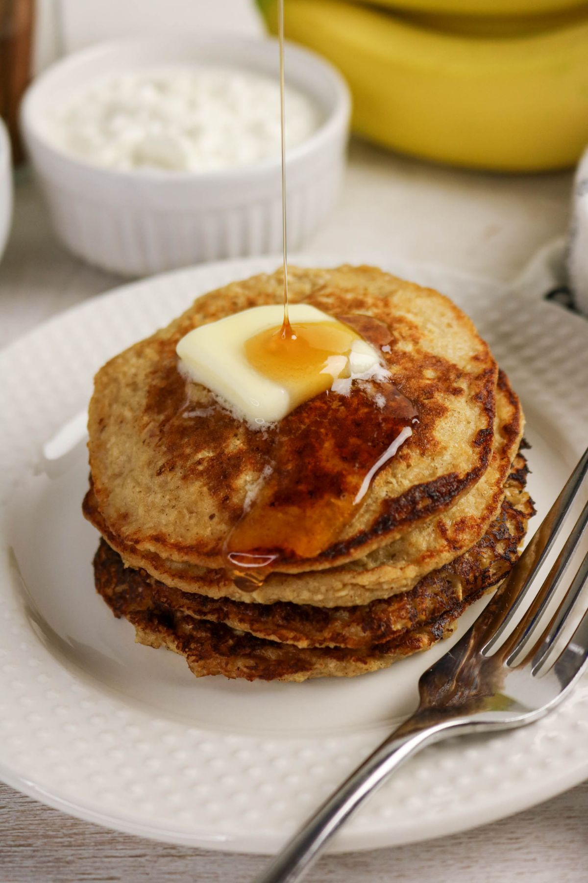 A stack of pancakes on a white plate with a pat of butter on top and syrup being poured over them, next to a fork.