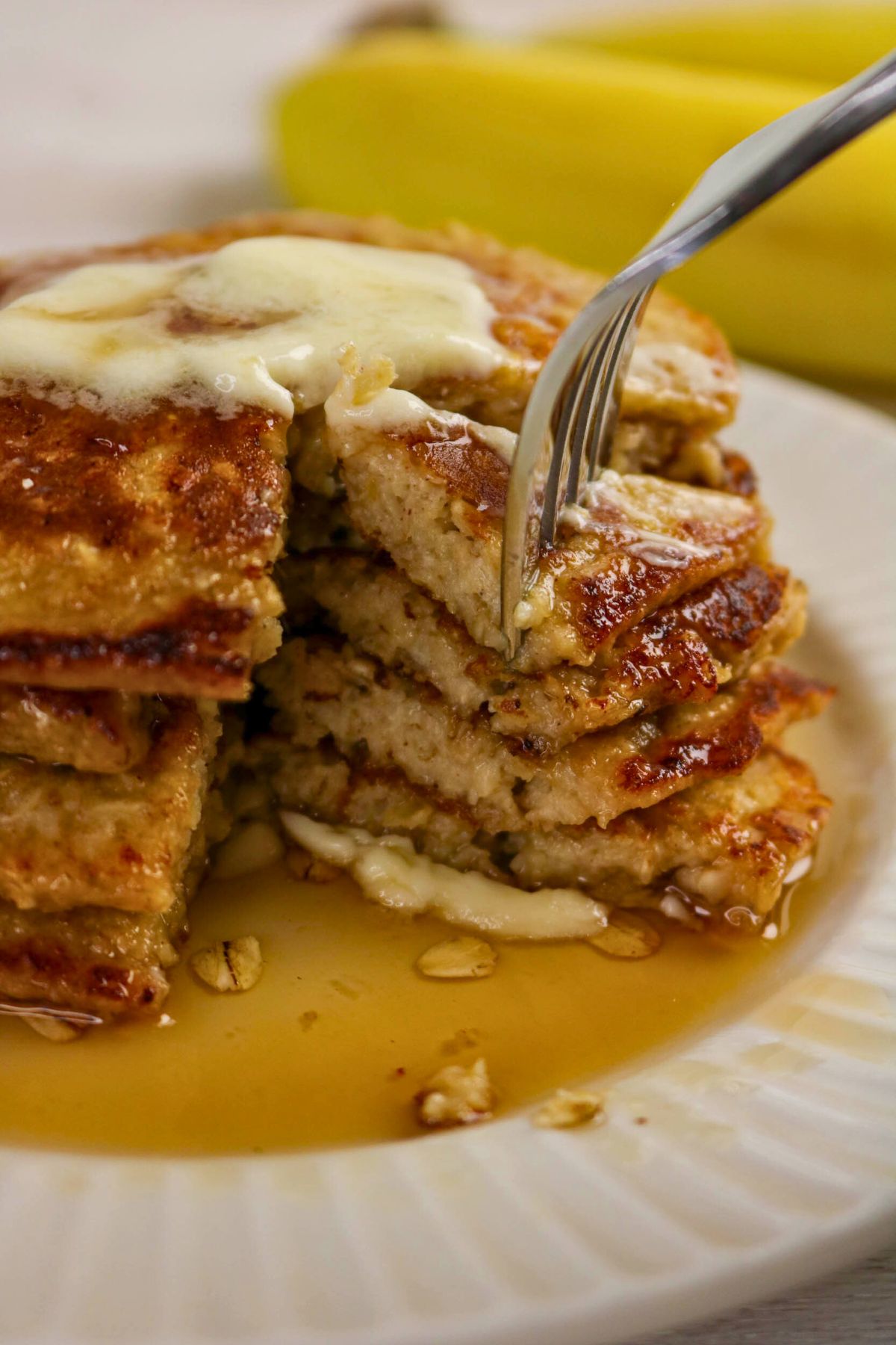 A stack of pancakes with melted butter and syrup on a white plate, with a fork cutting into them. Bananas are visible in the background.