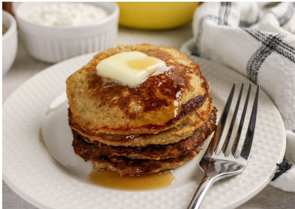 A stack of pancakes topped with butter and syrup sits on a white plate with a fork, next to a white napkin and small bowls in the background.