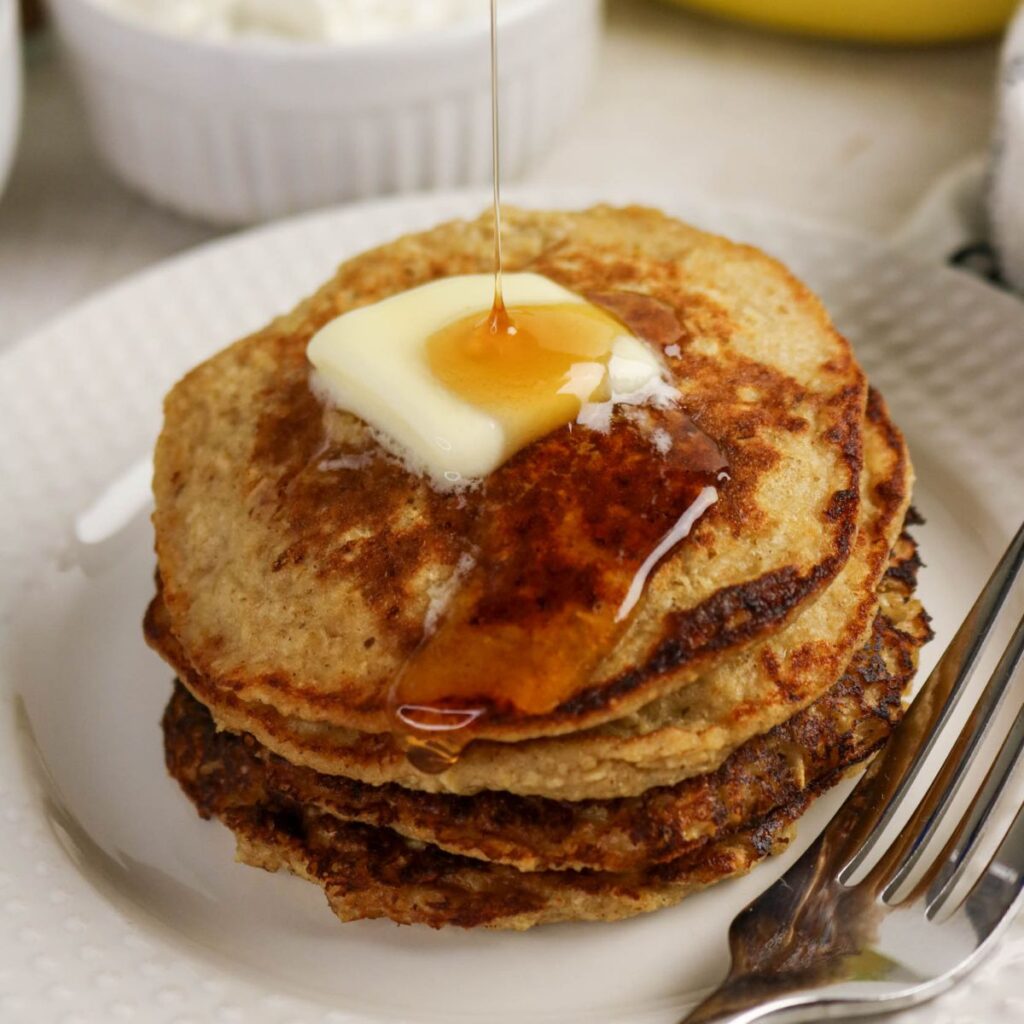 A stack of three pancakes topped with a pat of butter and syrup being poured over them, served on a white plate with a fork.