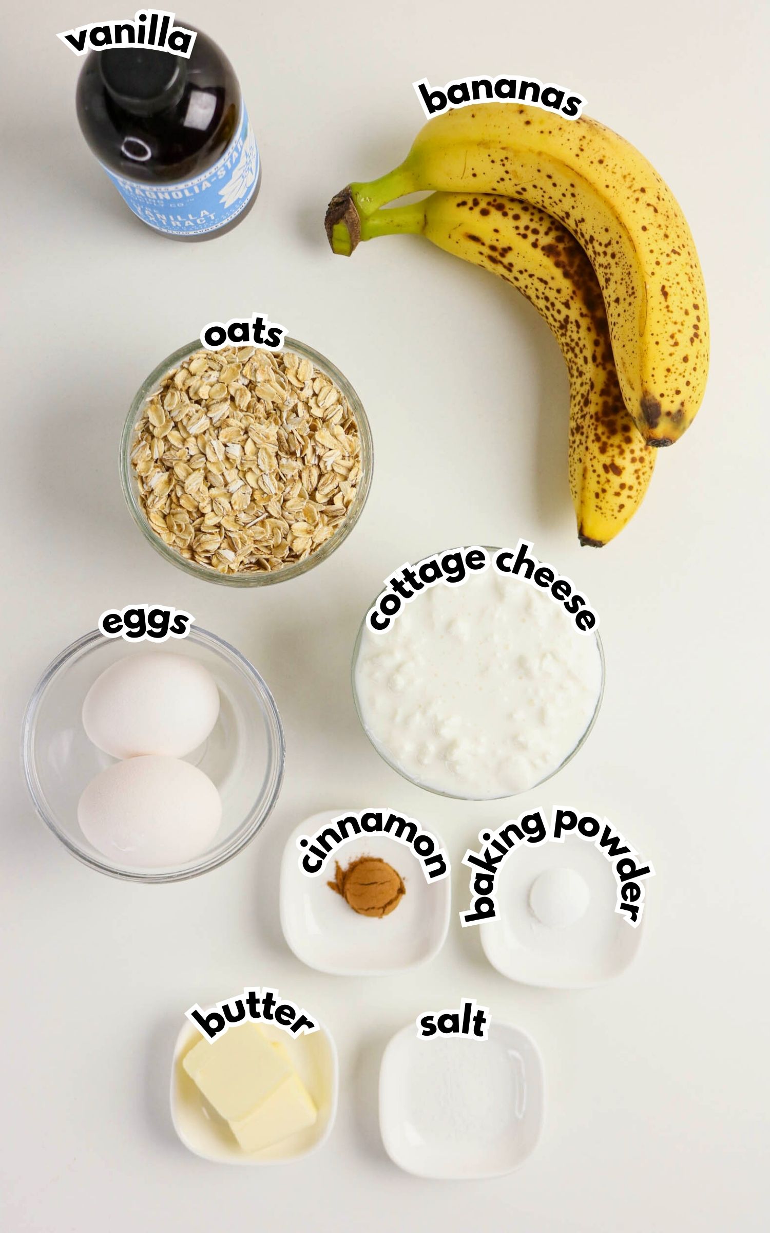 Top-down view of ingredients for a recipe: bananas, oats, cottage cheese, eggs, vanilla, butter, cinnamon, baking powder, and salt on a white surface.