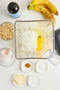 Overhead view of a blender with cottage cheese and eggs inside, surrounded by bananas, vanilla extract, oats, eggshells, cinnamon, butter, and salt on a white surface.