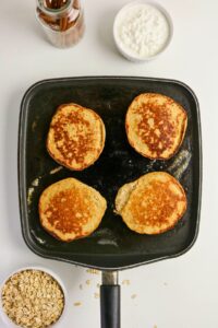 Four pancakes cooking in a square pan on a white surface, with bowls of oats, cottage cheese, and a jar of cinnamon sticks nearby.