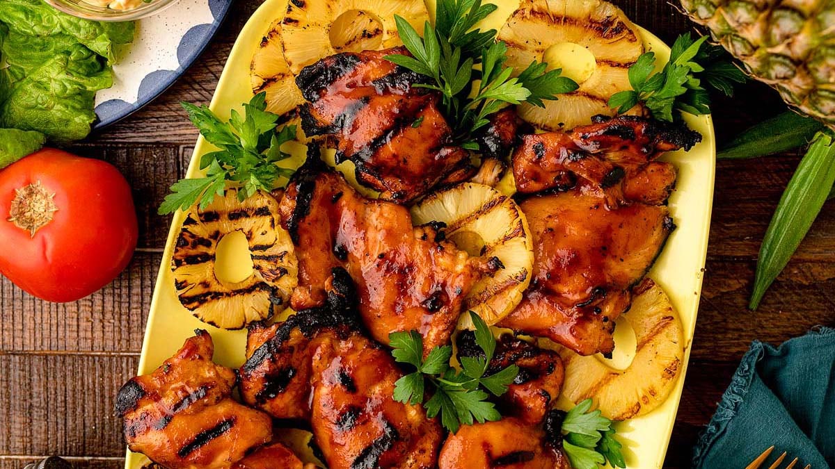 Grilled chicken thighs topped with barbecue sauce and garnished with parsley, served alongside grilled pineapple slices on a yellow platter.