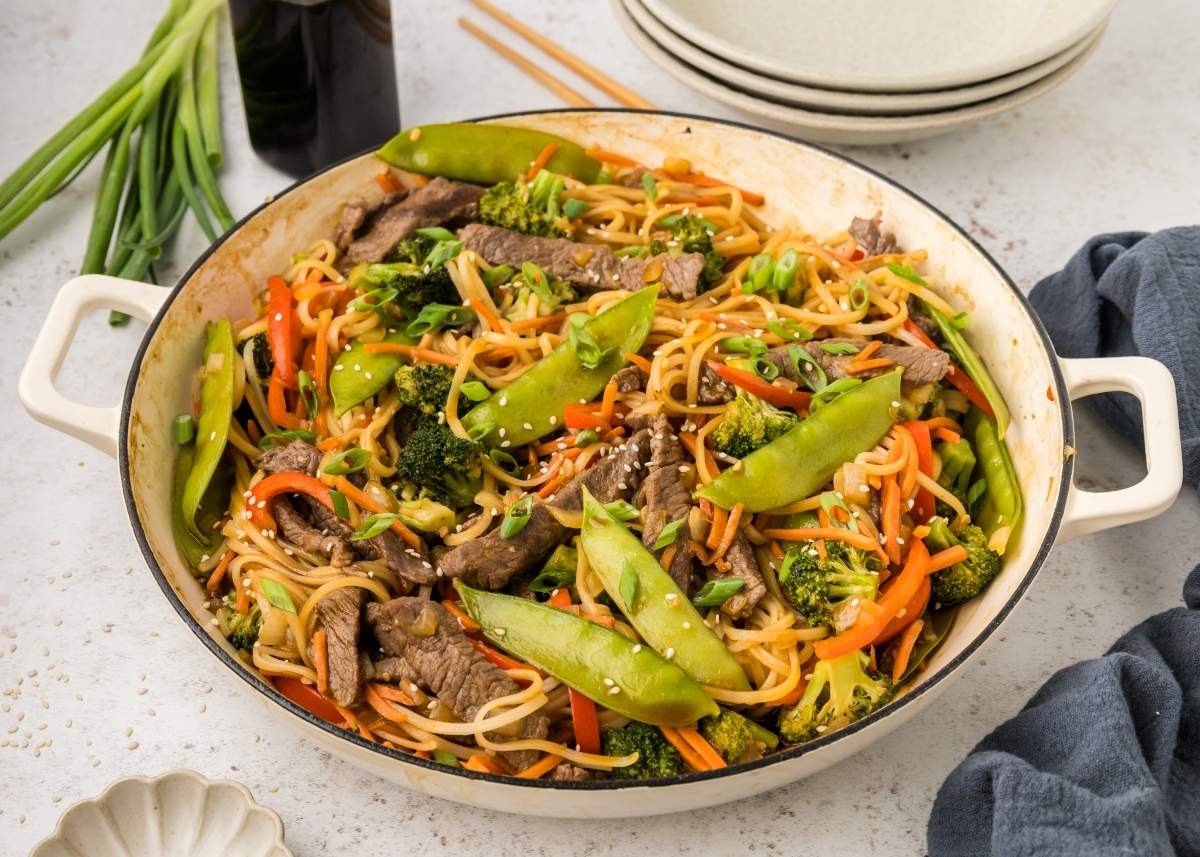 A pan filled with beef stir fry, noodles, snow peas, broccoli, carrots, and green onions, placed on a light countertop with plates and chopsticks nearby.