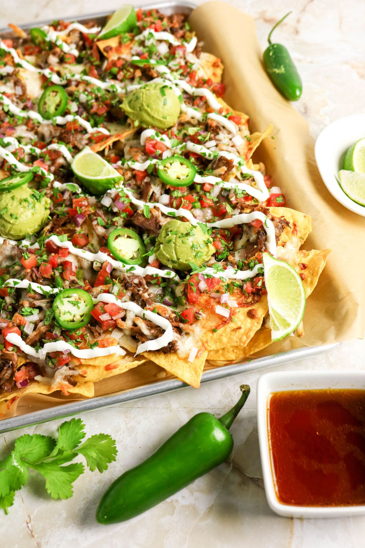 Sheet pan nachos topped with pulled meat, melted cheese, jalapeños, guacamole, sour cream, pico de gallo, and lime wedges, with salsa, cilantro, and jalapeños on the side.
