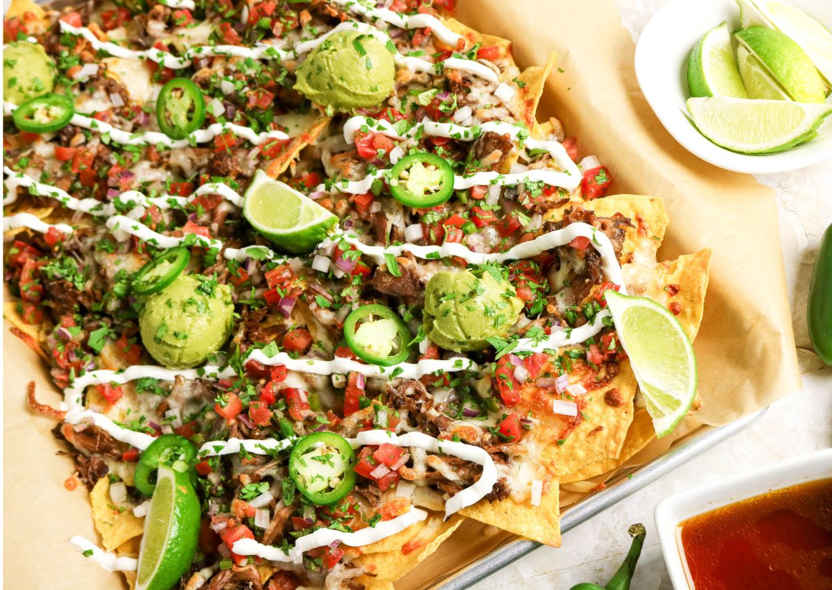 A tray of loaded nachos topped with shredded meat, guacamole, sliced jalapeños, pico de gallo, sour cream, and lime wedges.