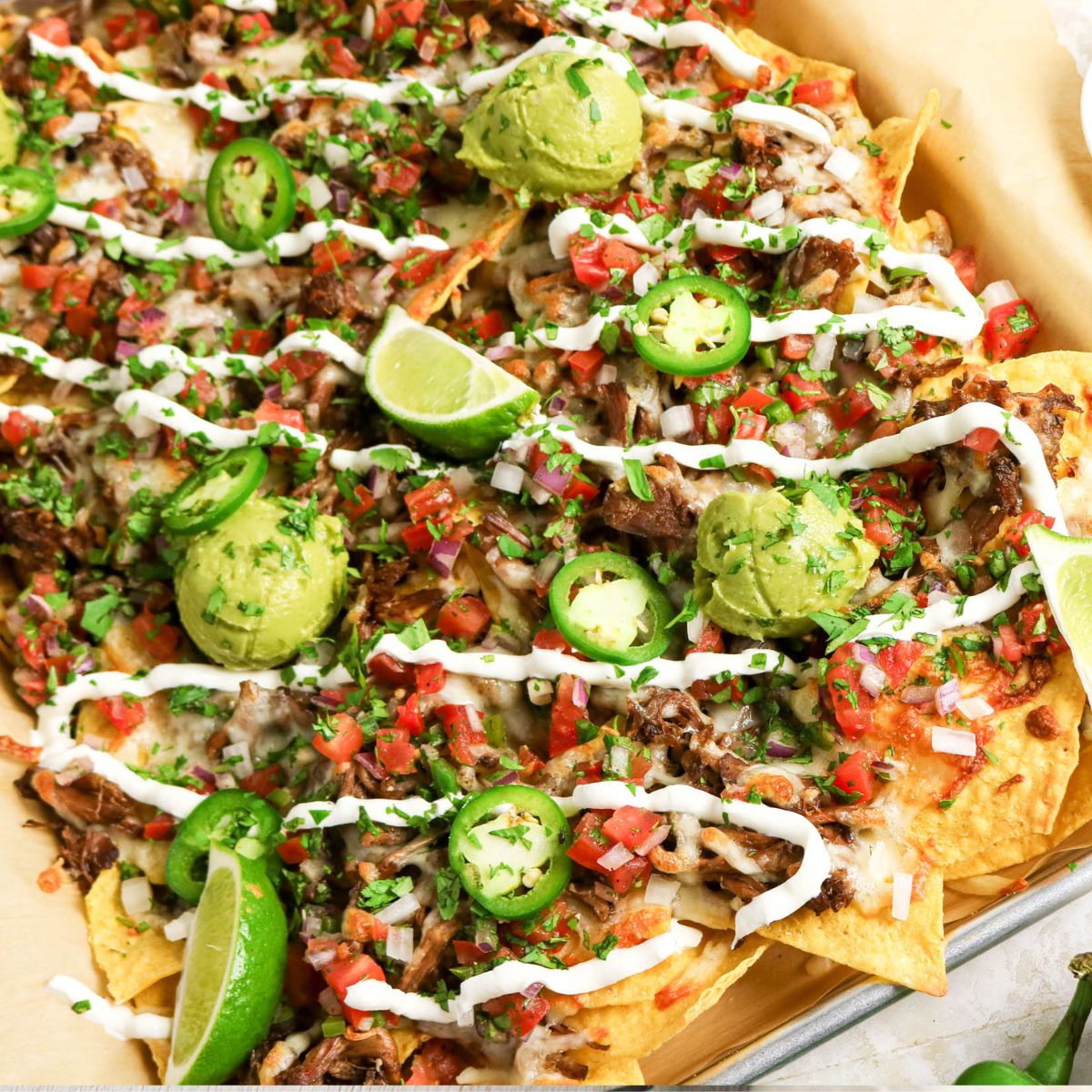 A tray of loaded nachos topped with shredded meat, pico de gallo, jalapeños, guacamole, lime wedges, melted cheese, and drizzled with sour cream.