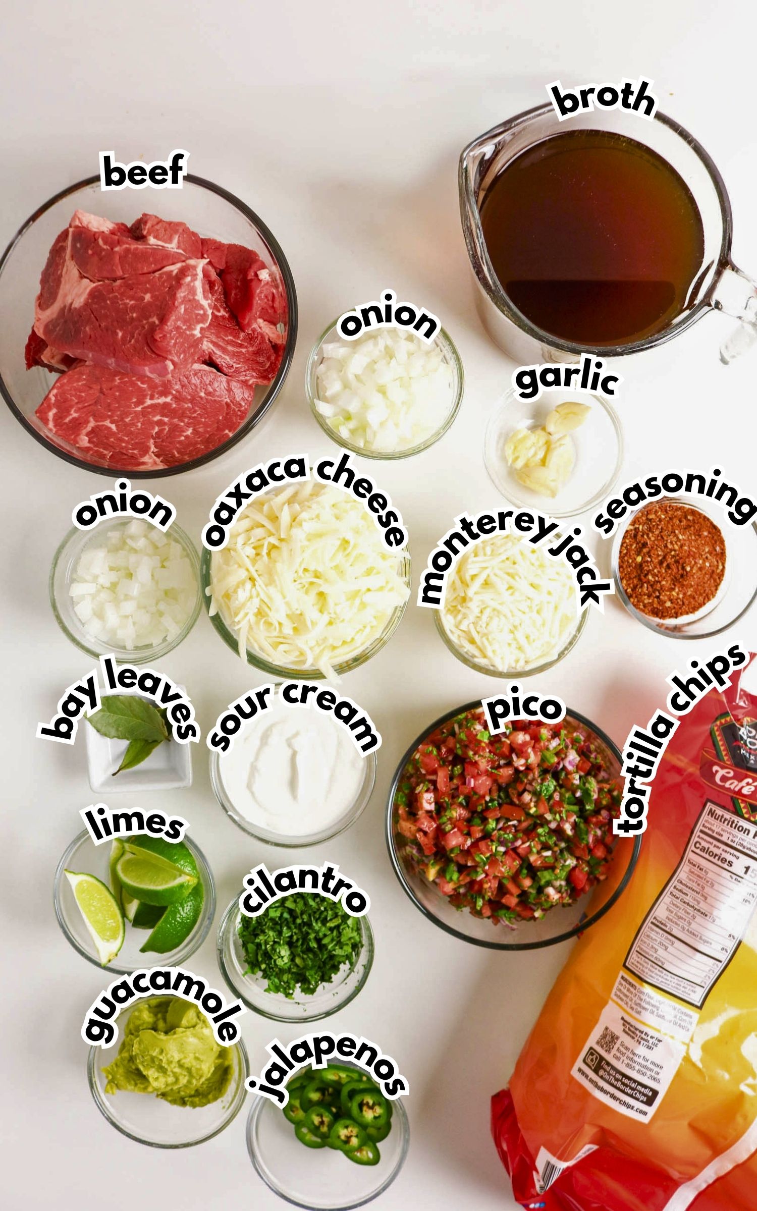 Various ingredients for a Mexican dish displayed, including raw beef, cheese, broth, diced onions, seasoning, pico de gallo, bay leaves, limes, cilantro, jalapenos, guacamole, sour cream, and tortilla chips.