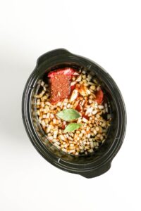 A black slow cooker filled with chopped onions, tomato paste, spices, bay leaves, and other ingredients before cooking.