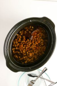 A slow cooker filled with diced meat in a dark sauce, next to a glass bowl containing metal tongs and forks on a white surface.
