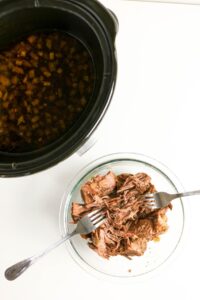 A slow cooker with broth next to a glass bowl of shredded beef being pulled apart with two forks on a white surface.