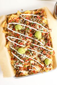 A tray of loaded nachos topped with melted cheese, pico de gallo, guacamole, and drizzled sour cream on parchment paper.