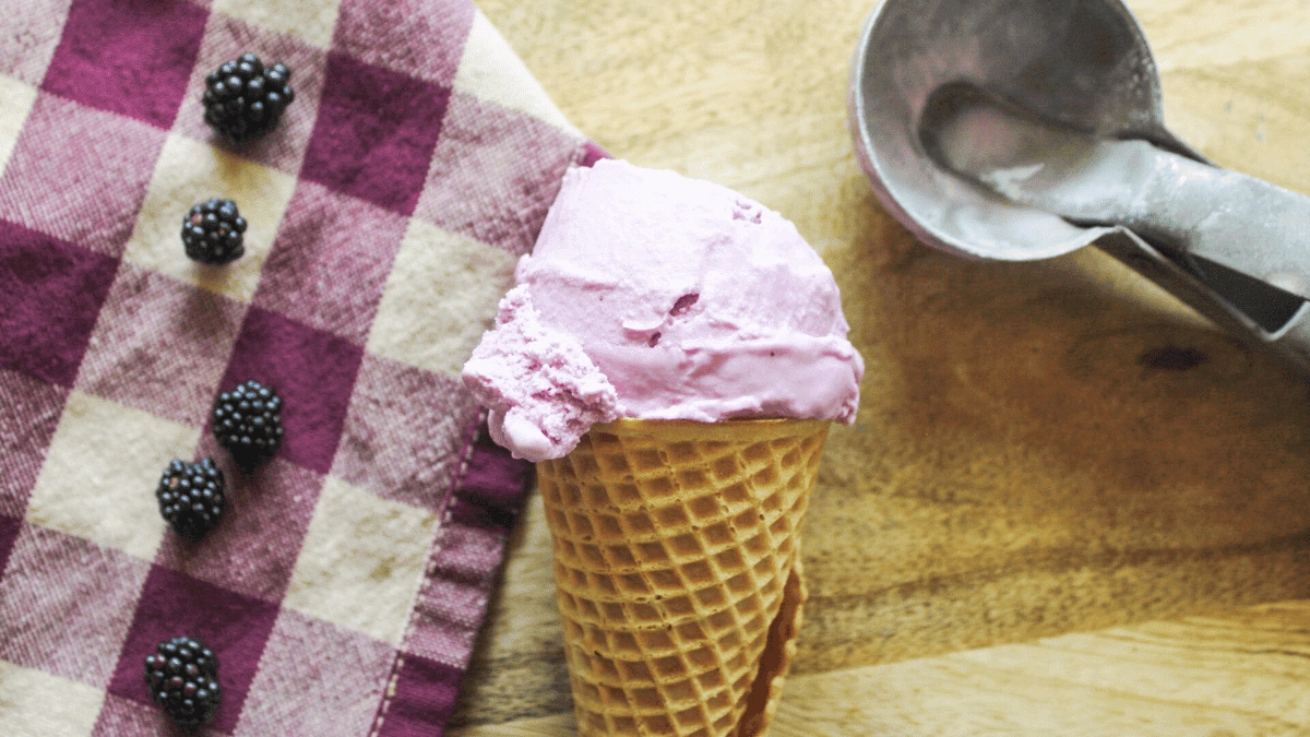 A waffle cone with a scoop of purple ice cream sits next to a metal ice cream scooper, blackberries, and a checkered cloth on a wooden surface.