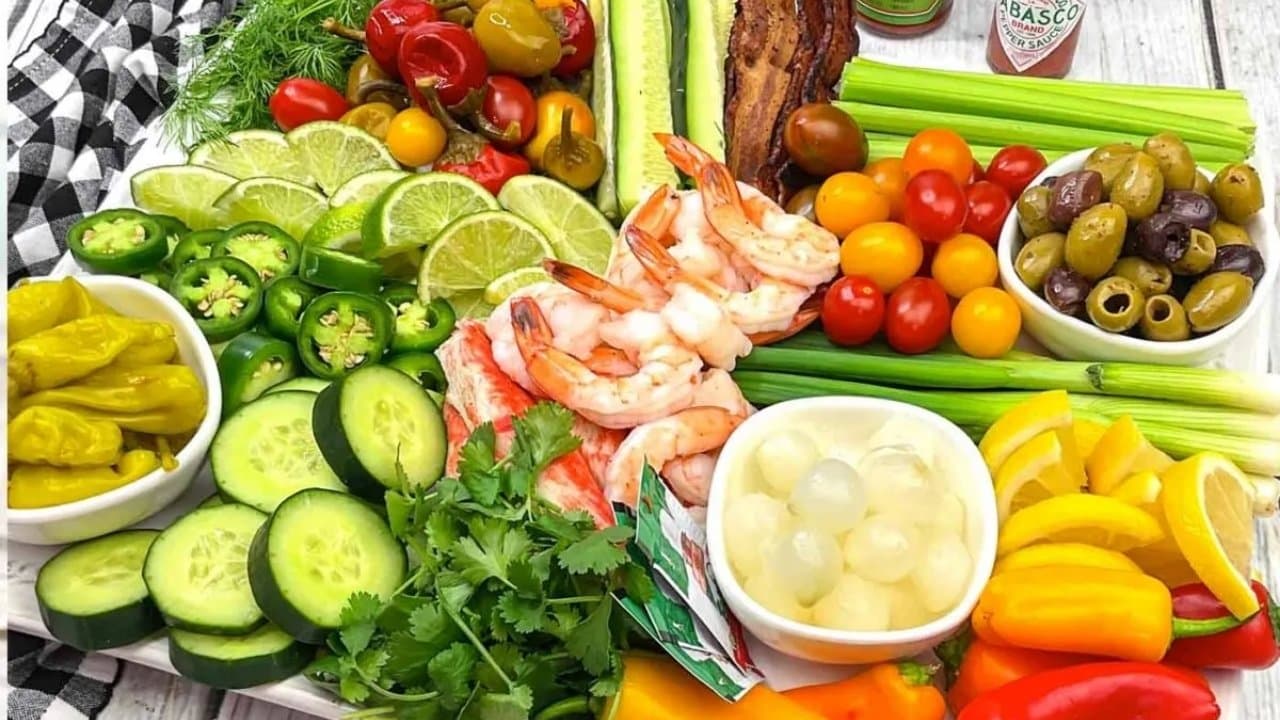 A platter with shrimp, sliced cucumbers, bell peppers, lime wedges, cherry tomatoes, olives, celery sticks, jalapeños, pepperoncini, cocktail onions, and bacon strips.