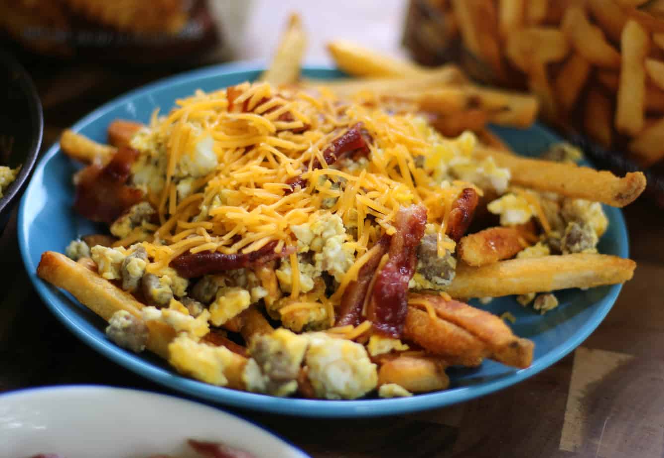 A blue plate of French fries topped with scrambled eggs, bacon, sausage, and shredded cheddar cheese.