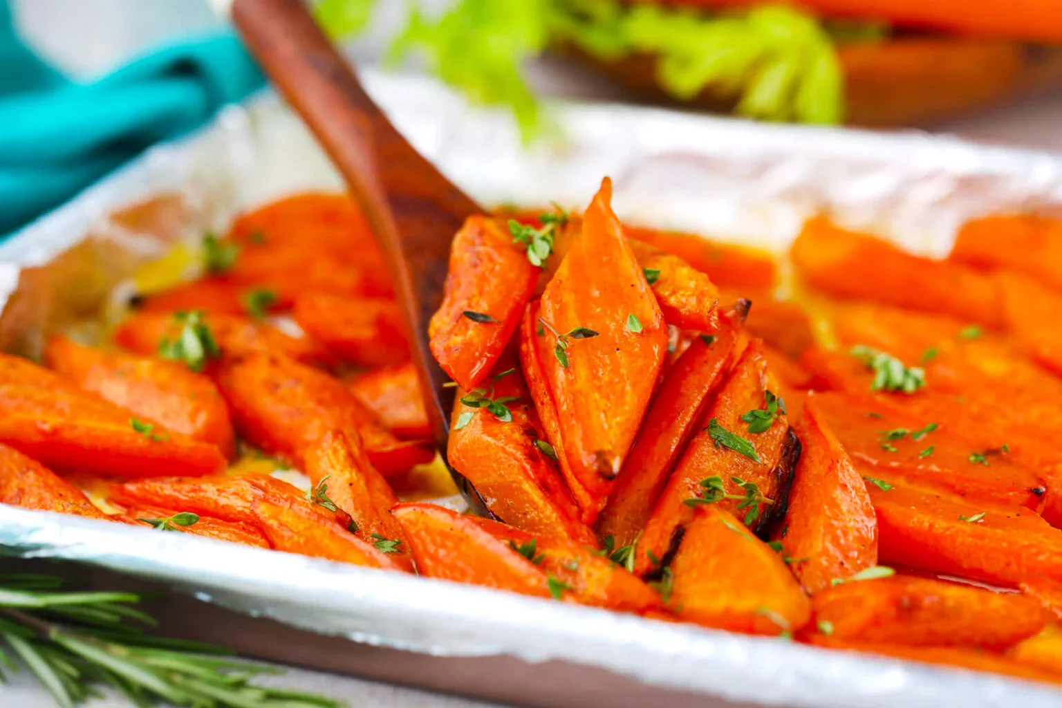 Roasted carrot slices garnished with chopped herbs on a foil-lined baking tray, with a wooden spoon lifting some of the carrots.