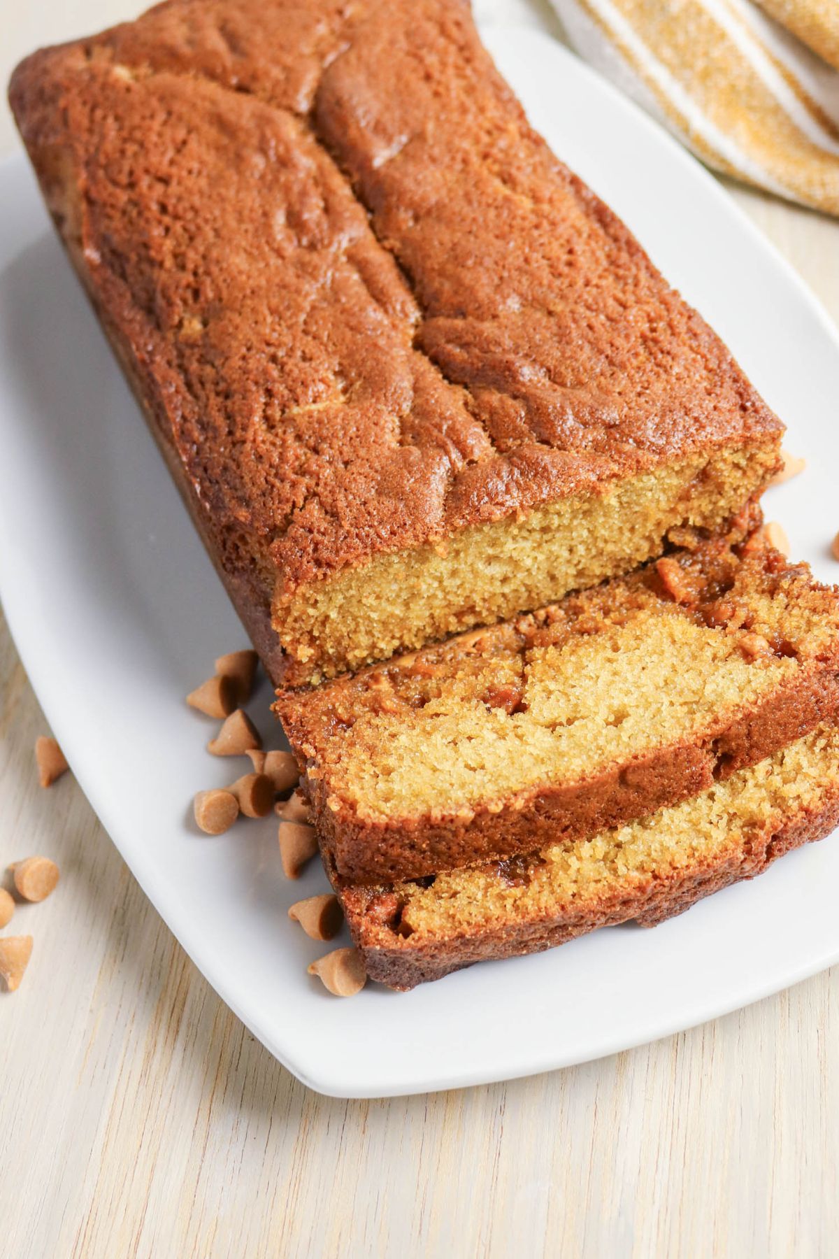 A loaf of golden-brown bread with two slices cut, displayed on a white plate with butterscotch chips scattered around.