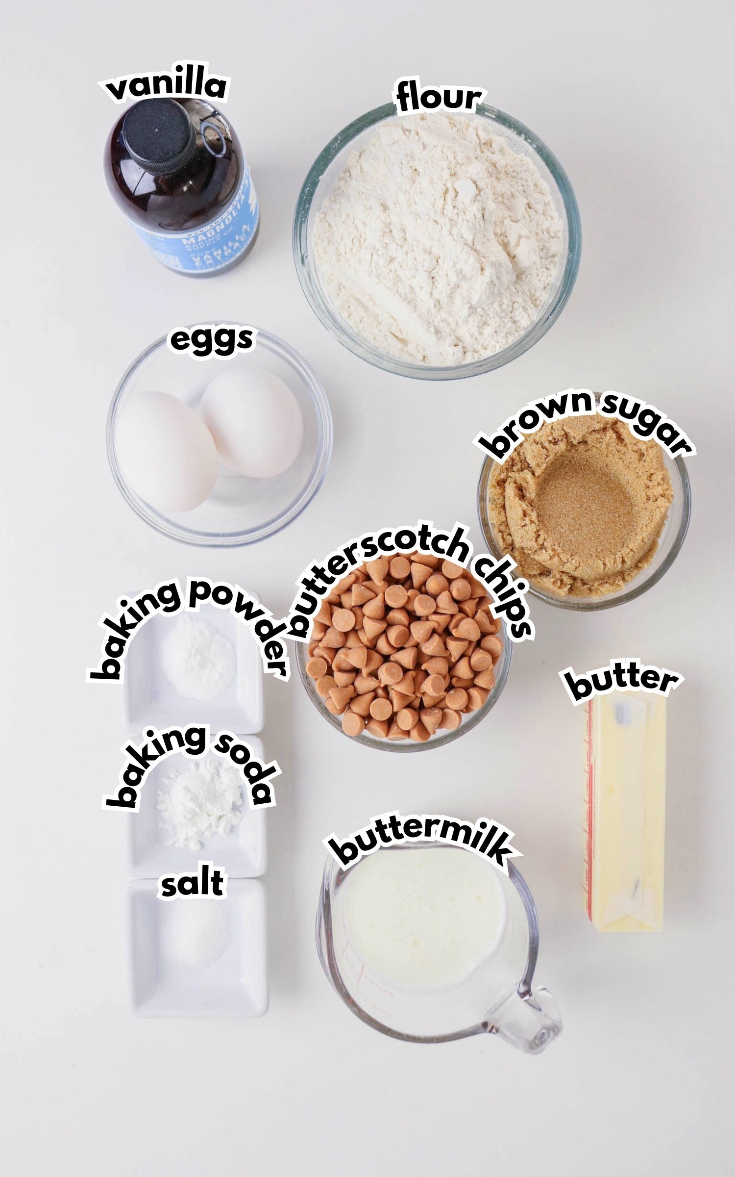 Top-down view of labeled baking ingredients: vanilla, flour, eggs, brown sugar, butter, butterscotch chips, buttermilk, salt, baking soda, and baking powder on a white surface.