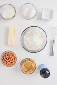 Baking ingredients arranged on a white surface, including flour, eggs, butter, brown sugar, butterscotch chips, vanilla extract, milk, salt, and a whisk.