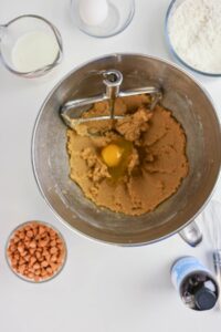 A mixing bowl with cookie dough and a cracked egg, surrounded by butterscotch chips, flour, an egg, milk, vanilla extract, and a whisk on a white surface.