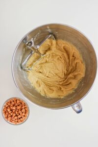 A mixing bowl with cookie dough and a metal beater next to a small bowl filled with butterscotch chips on a white surface.