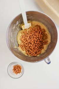 A mixing bowl contains cookie dough with butterscotch chips and a spatula; a small glass bowl of extra butterscotch chips is nearby.