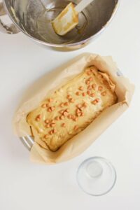 A loaf pan lined with parchment paper holds unbaked batter topped with butterscotch chips. A spatula rests in a metal mixing bowl nearby, with an empty glass bowl beside them.