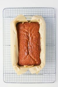 A loaf of baked bread in a parchment-lined loaf pan, resting on a metal cooling rack.