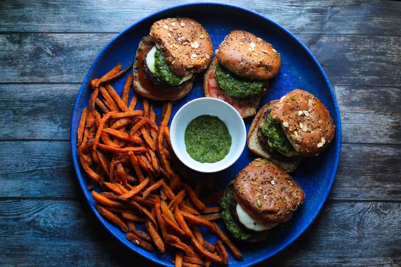 A blue plate with three veggie burgers, sweet potato fries, and a small bowl of green dipping sauce on a wooden table.