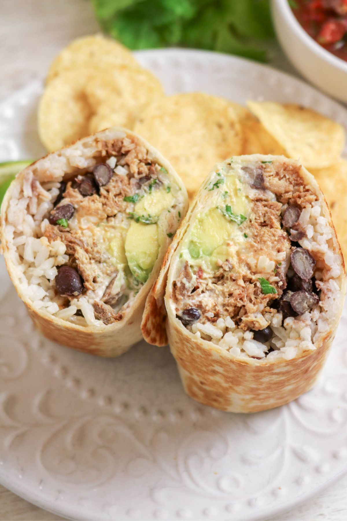 A cut Carnitas Burrito on a plate reveals rice, black beans, tender shredded meat, avocado, and sauce inside; tortilla chips are in the background.