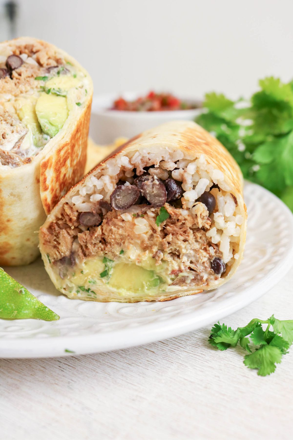 A burrito cut in half on a white plate, showing rice, black beans, shredded meat, avocado, and cilantro inside. Lime and cilantro garnish are nearby.